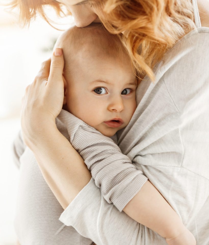 Portrait of sweet little child looking in the camera with interested expression while mother tenderly snuggle palming and kissing her newborn baby